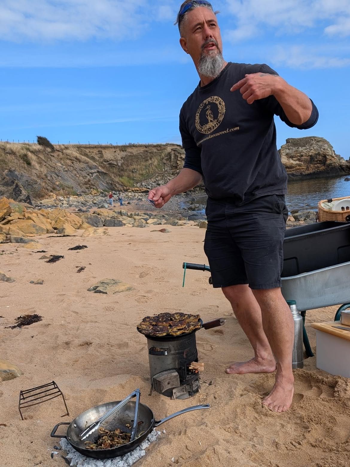 Jayson at the beach kitchen on the sand, a pan on the stove and the shore behind