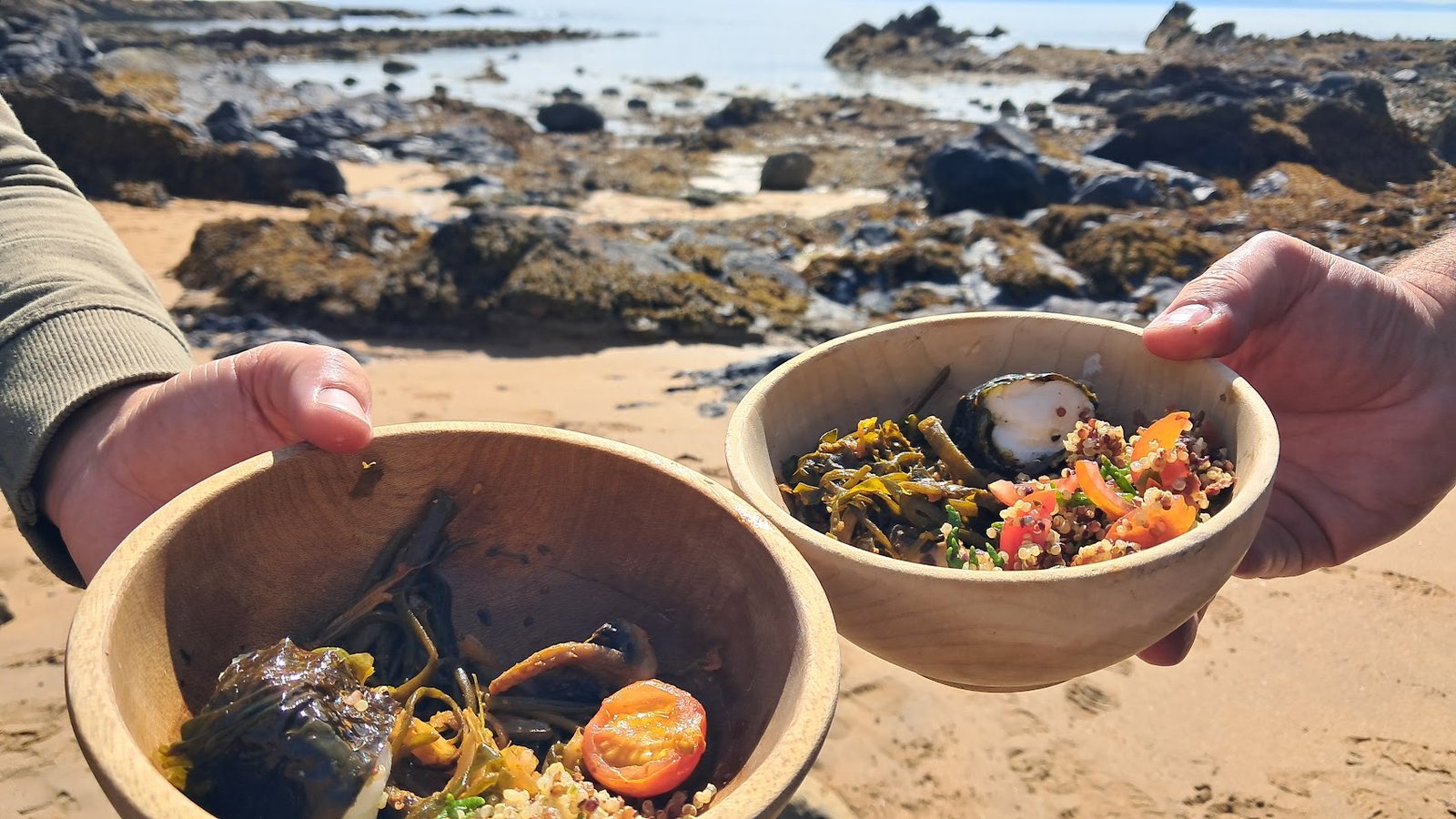 Two hands holding out wooden bowls of seaweed, grains and tomatoes on the beach