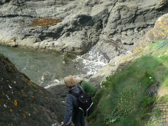Two walkers descending steep rock towards a foaming inlet on the Elie Chain Walk, chain in hand