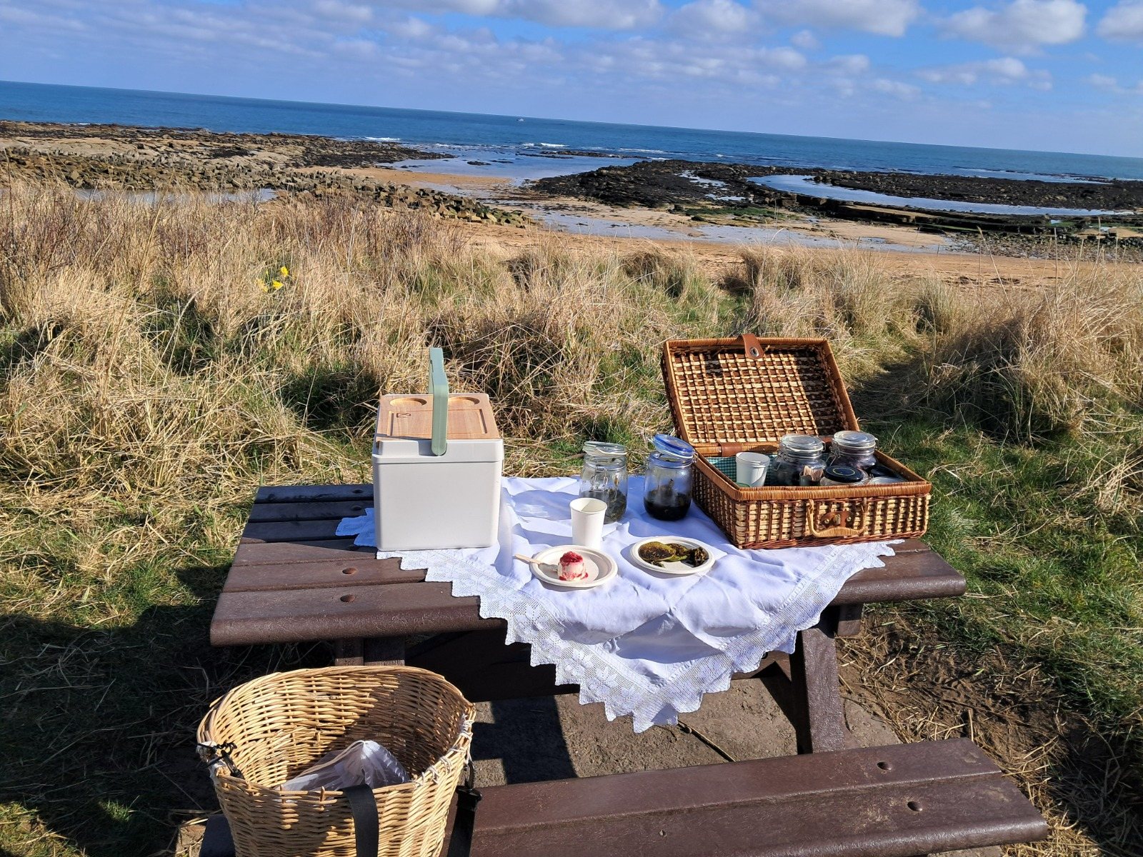 A picnic table laid with a cloth, baskets and jars above the shore