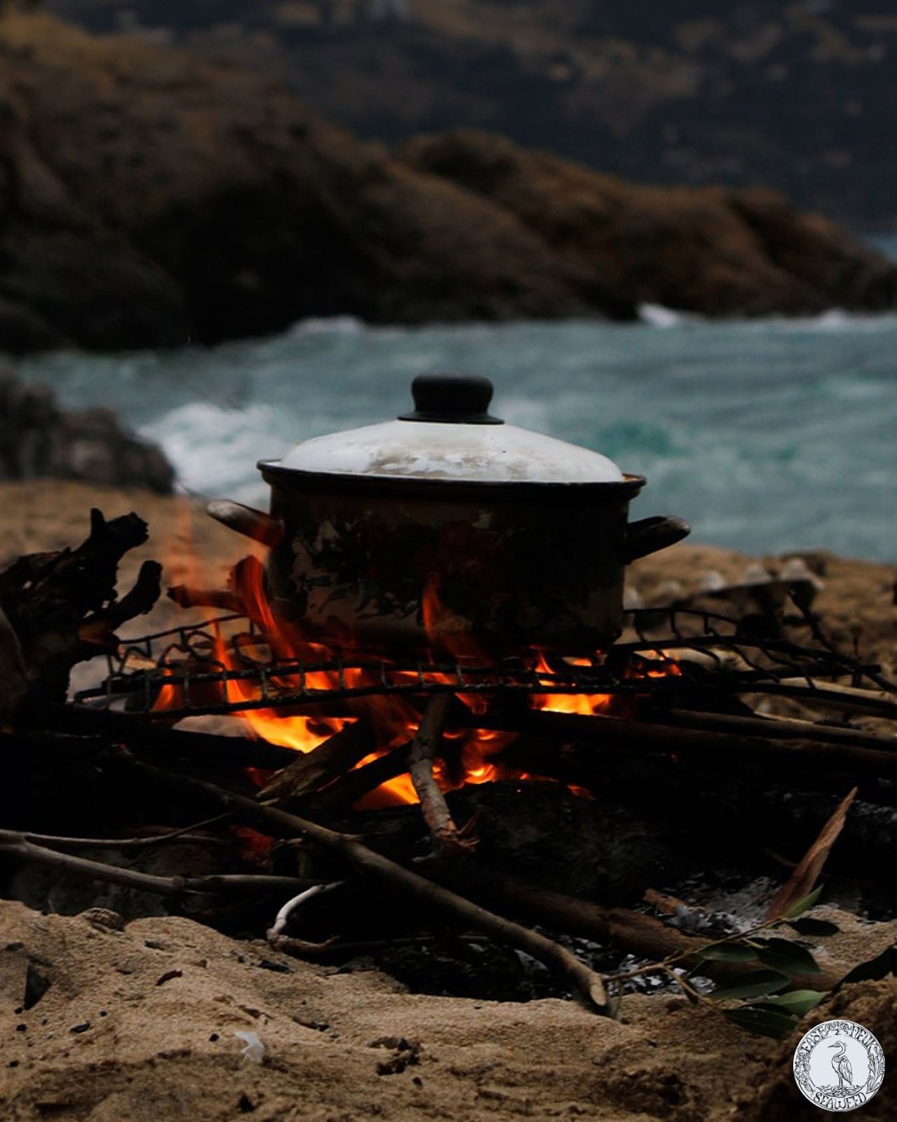 A pot on an open fire on the shore at dusk, waves breaking behind