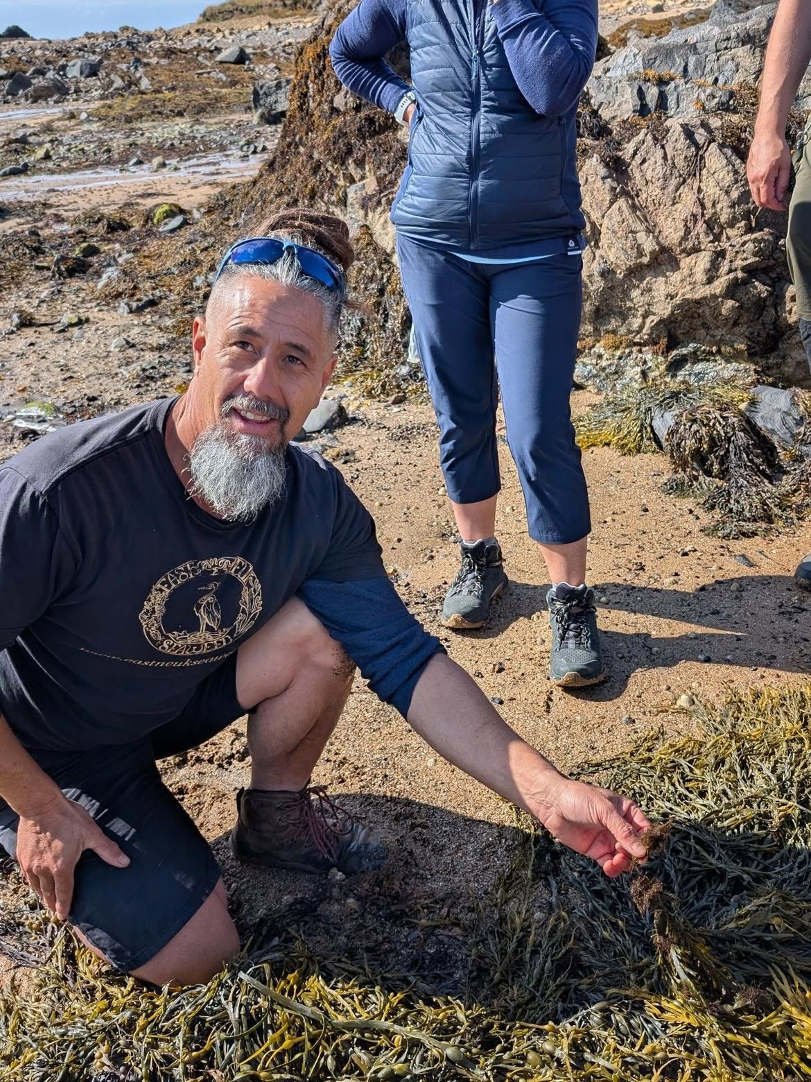 Jayson kneeling on the wrack-covered shore, pointing out a seaweed to guests