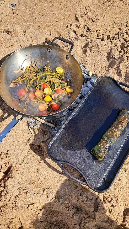 A wok of vegetables and seaweed over a fire on the sand, a wrapped seaweed parcel on a griddle beside it