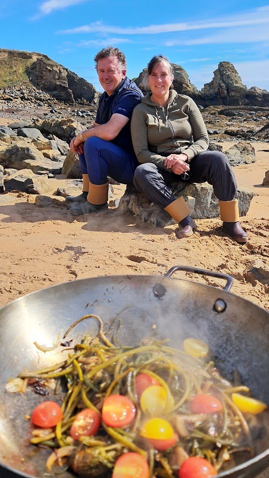 Two guests sitting on the rocks on the beach, a steaming wok of seaweed and tomatoes in the foreground